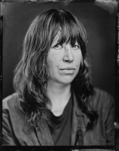 Black and white portrait of a woman with mid-length wavy hair and bangs. The edges of the photo are distressed to create the effect of an older photo