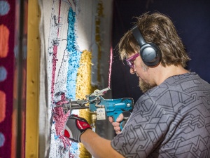 Man wearing glasses and headphones uses a tufting gun on a large frame