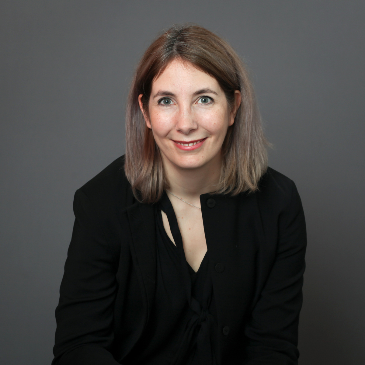 Headshot of a person with mid-length hair in black v-neck shirt against dark graynbackground.