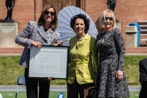 Three women standing side by side, the farthest to the left, Jennifer Gilbert in black pants and plaid boucle blazer, is holding a framed degree. In the middle, Artis Lane in a chartreuse blazer and brown pants holds a white sun parasol behind her. On the right stands Susan R. Ewing in a black and white patterned long sleeve dress. Brick building and bronze sculptures in the far background.