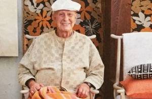Waist up portrait of an older man with light cardigan and white cap in front of a floral patterned wall.