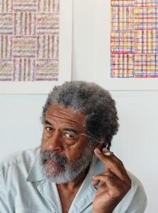 A shoulder-up headshot of a man with grey hair and beard holding a cigarette between his first two fingers. Behind him are two geometric, abstract artworks, the lower corners of each is visible over the man's head.