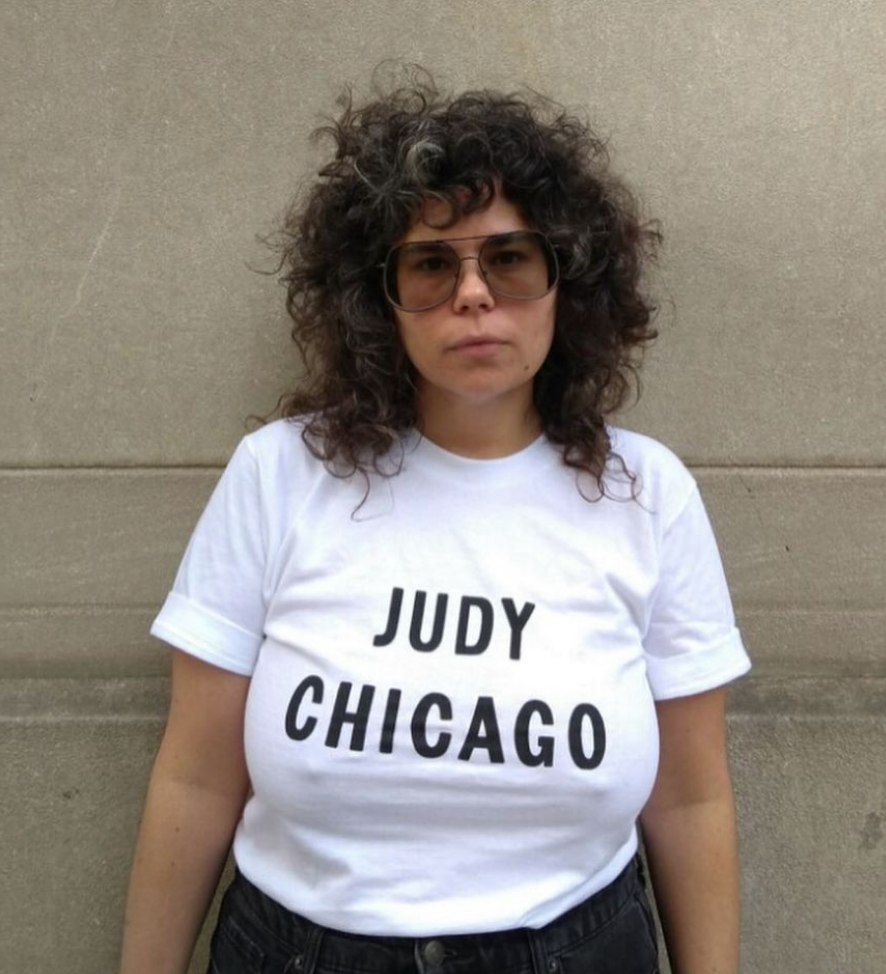 Portrait of a curly-haired person from the waist up wearing a white t-shirt. Bold, black text on the front of their shirt reads, "Judy Chicago".