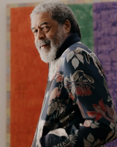 Portrait of artist McArthur Binion standing in front of a red, green, and purple vertically-striped artwork. He stands with his left side facing the camera, with his left hand in his pants pocket and face turned towards the camera.