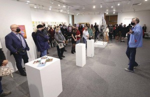 A group of people in a gallery space with white walls and gray flooring listen to a person in blue shirt and jeans present. Several white pedestals with art objects stand in between the group of listeners and the presenter.
