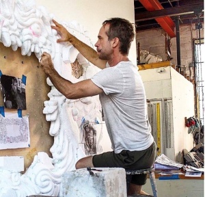 Photo of artist Chris Schanck at work in his studio. A person in a while shirt and black shorts stis on a metal stool sculpting a large white frame made of organic shapes. THe frame is resting on the floor and slightly angled back to lean on a wall that has a few sketches taped up for reference.