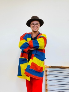 A person stands with their arms crossed in front of a white wall. They are wearing a brown, wide-brimmed hat, glasses, a colorful cardigan, and red pants.