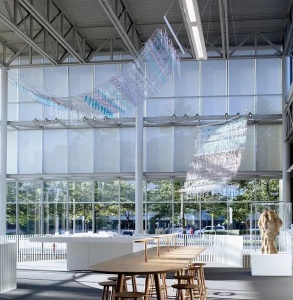 An art installation is suspended from exposed beam ceiling in a large room with wall-to-wall windows. Two sheet-like installations made up of various medical waste objects hang over a long wooden table surrounded by stools.