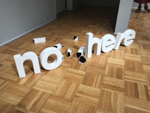 A ceramic sculpture sits on wooden parquet flooring in a gallery space with grey walls. The sculpture is made up of multiple ceramics pieces in white, that form the words "no here" in three-dimensional Helvetica font. Several smaller white and black ceramic pieces that look like a broken "w" are scattered in between the letters "no" and "here".