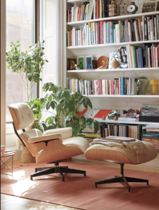 An Eames Lounge Chair with Ottoman viewed from the side in front of a large built-in white bookcase. The chair is upholstered in white leather with a light, warm wood. The base in black.To the left, behind the chair is a window and tall green plant.