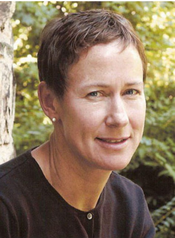 A headshot of a person with short brown hair against a background of green leaves and trees