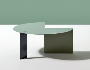 A muted green steel coffe table against a white floor and green wall. The table is round with an angular negative space creating a storage nook and support. An additional flat rectangular leg is a contrasting color.