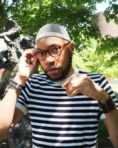 A person with glasses and a beard. They are wearing a black and white horizontally-striped t-shirt and a light-colored mesh cap. Their right hand reaches up to grasp the side of their glasses. Their left hand in lifted up naturally in front of their chest. In the background is green foliage and the edge of a stone sculpture.