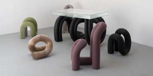 Various sculptural furniture pieces are arranged on a grey floor with white background. The chairs, ottomans, and talbe are made up of curving cylinders, in several colors and materials.