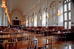 An angled view in a long historic dining hall. Wooden tables and chairs are lined up on the left and right side. The ceiling is arched and the walls feature many large arched windows with unique leaded glass designs.