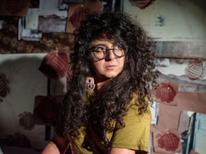 A chest-up portrait of a person with long curly hair. They are wearing an olive short sleeve shirt and unique eyeglasses with frames made of coiled wire. Their long hair is pulled forward of their shoulders and they are looking over their left shoulder looking out of frame.