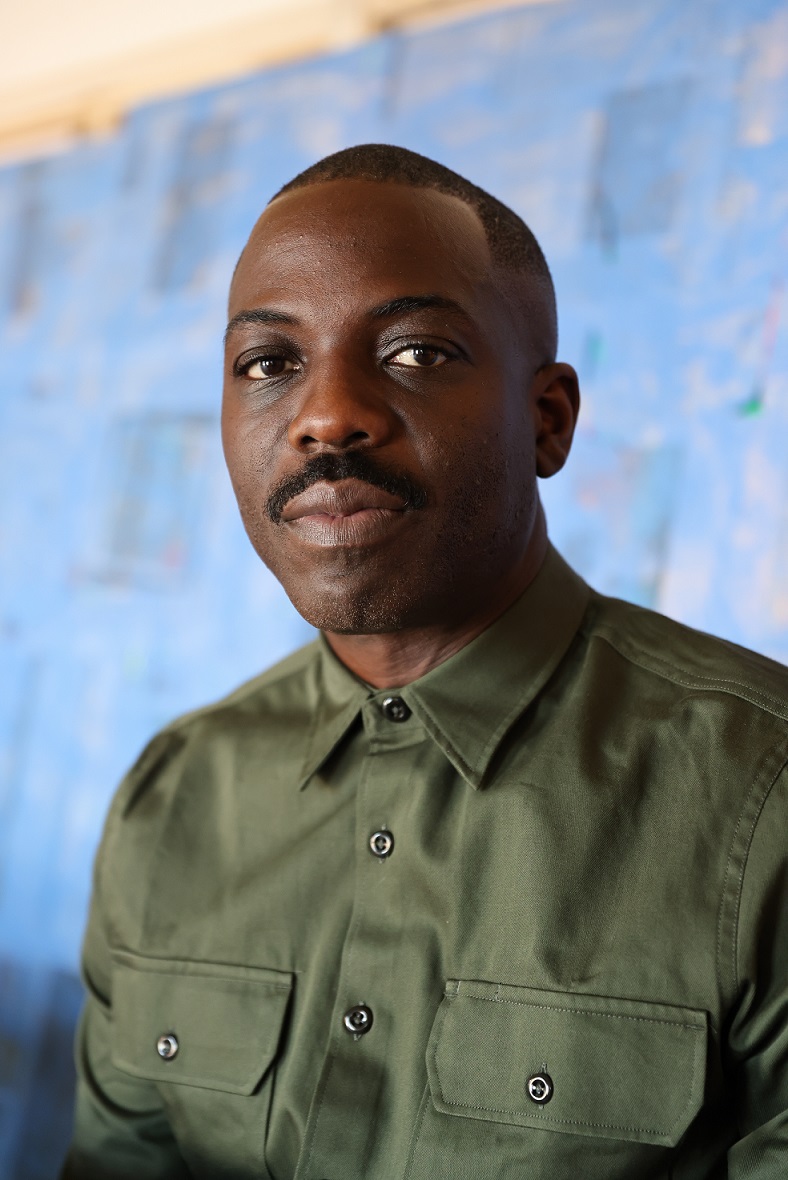 A chest up portrait of a personwith close-shaven hair and mustache wearing a green button-down shirt against a blue background.