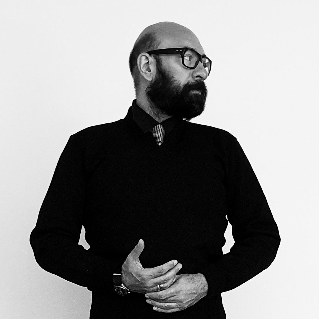 A black and white portrait of a person with a beard and thick-framed glasses wearing a v-neck sweather, tie, and collared shirt. Theiy are clasping their hands in front of their waist, a watch on their right wrist.