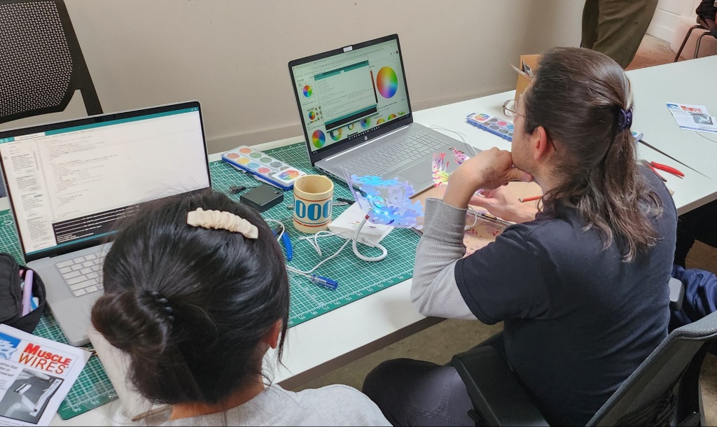 Two students looking at coding program on their laptops at a grey table. Small electronics equipment is scattered on the surface near their laptops.