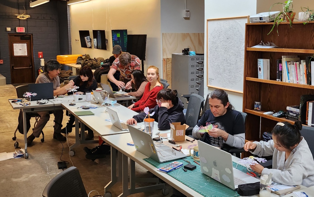 Group of students sit along one side of a long l-shaped group of tables. Laptops and small electronics devices are gathered in front of each student.