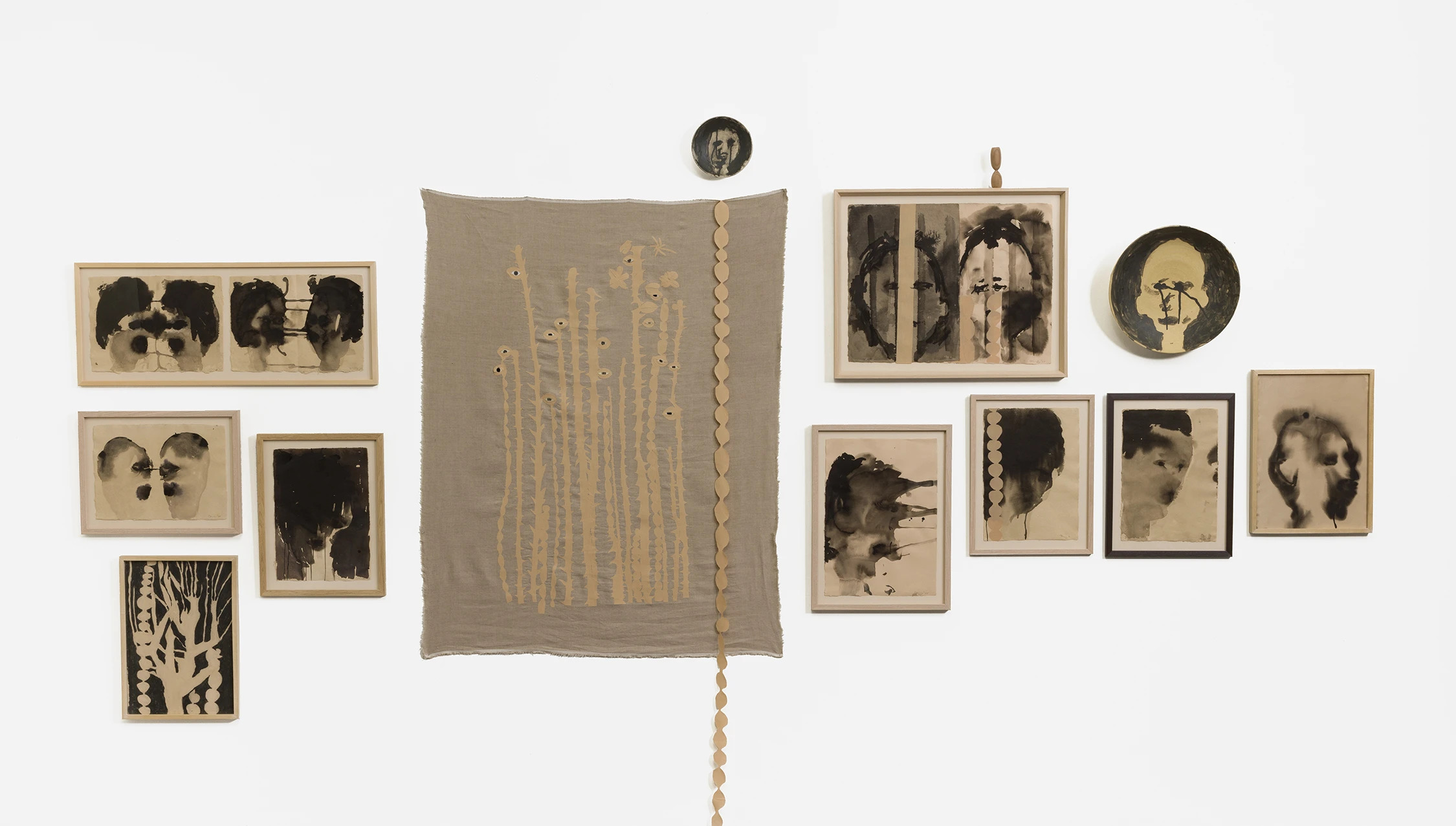 A monochrome art installation featuring a central fabric piece with botanical-like forms with eyes, flanked by framed expressive ink portraits and botanical works. 