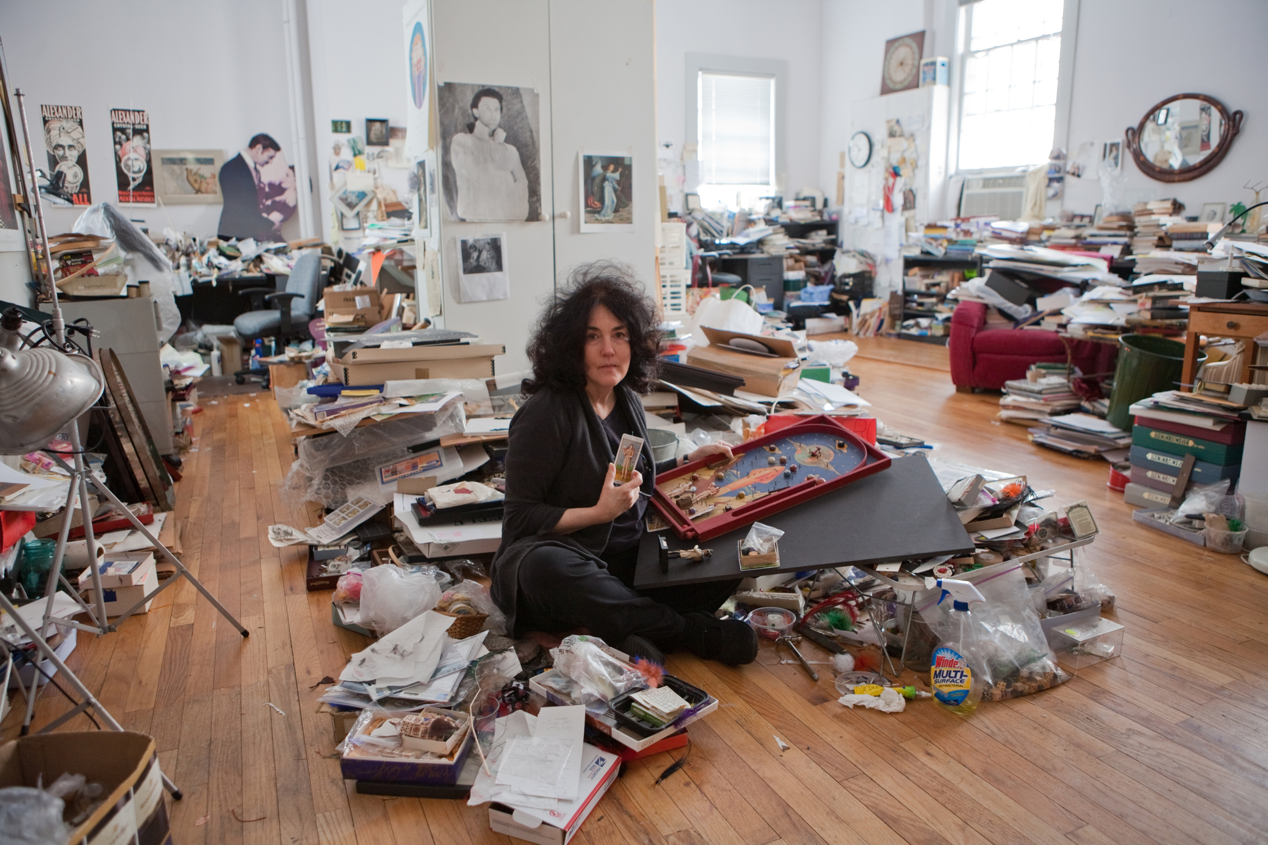 A person wearing a black sweater and pants sits cross-legged in the middle of a large room with wood flooring. The person is examining artwork and found objects gathering in large piles around them and clustered throughout the room. Piles of books and other ephemera fill their studio space. Artwork, posters, a mirror, a clock, etc. are pasted on the walls in the background. 