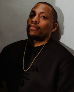 Headshot of a person with a beard and mustache wearing a black shirt and gold chain against a grey backdrop.