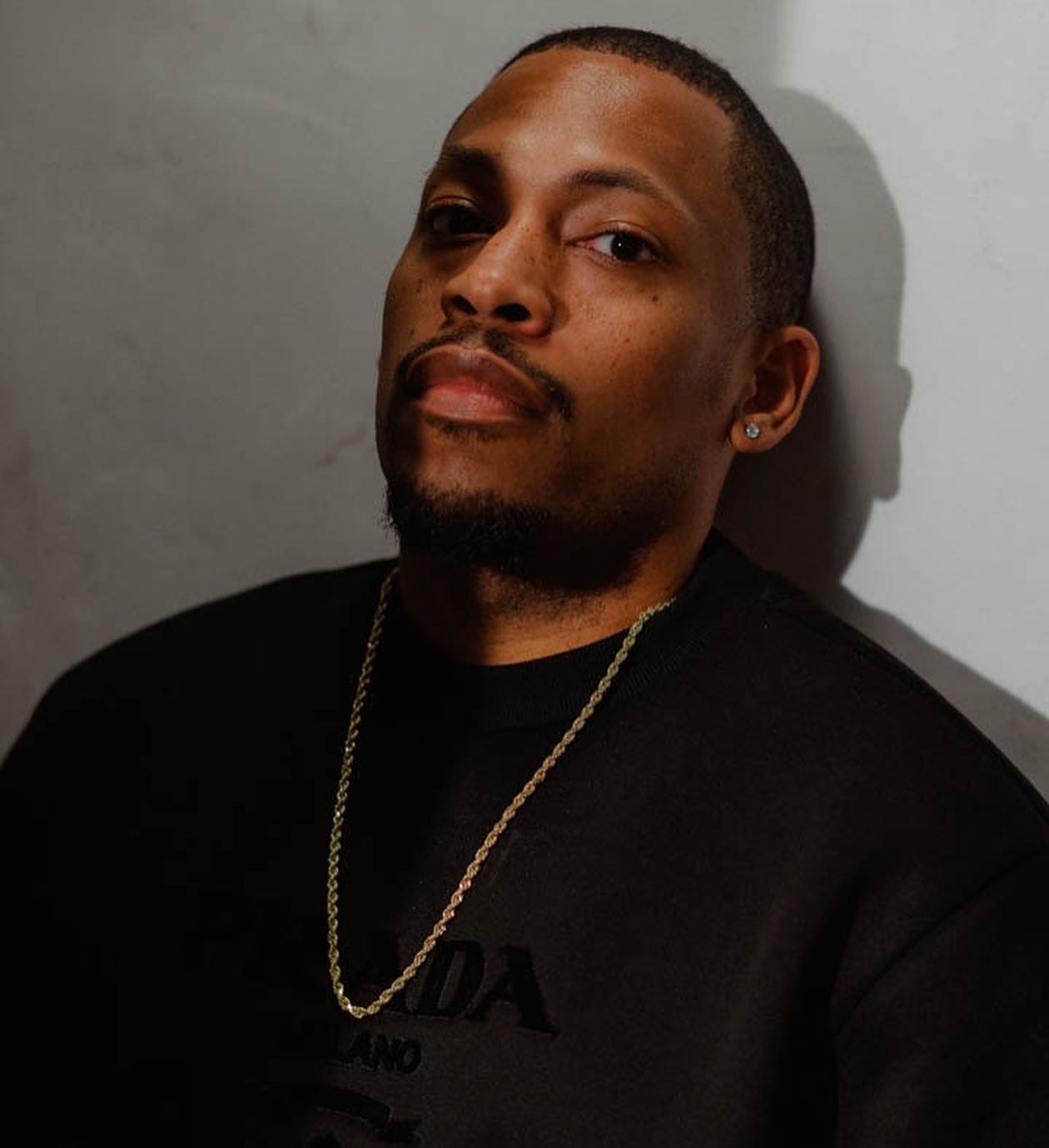 Headshot of a person with a beard and mustache wearing a black shirt and gold chain against a grey backdrop.