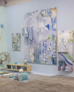 An art gallery room featuring abstract, colorful paintings in shades of blue, green, and purple. The walls display multiple canvases with intricate patterns and textures. A light wooden shelf holds assorted decorative items, including vases and folded fabric. The floor has a small rug and a few large spheres. Overall, the setting is vibrant and contemporary.