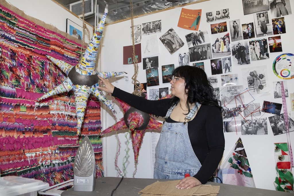 A person in a denim apron stands in an art-filled studio, touching a large, colorful star-shaped piñata-like sculpture suspended from the ceiling. The wall is covered with numerous black-and-white and color photos photos, and other decorations. An iron and burlap material are on the ironing table in foreground.