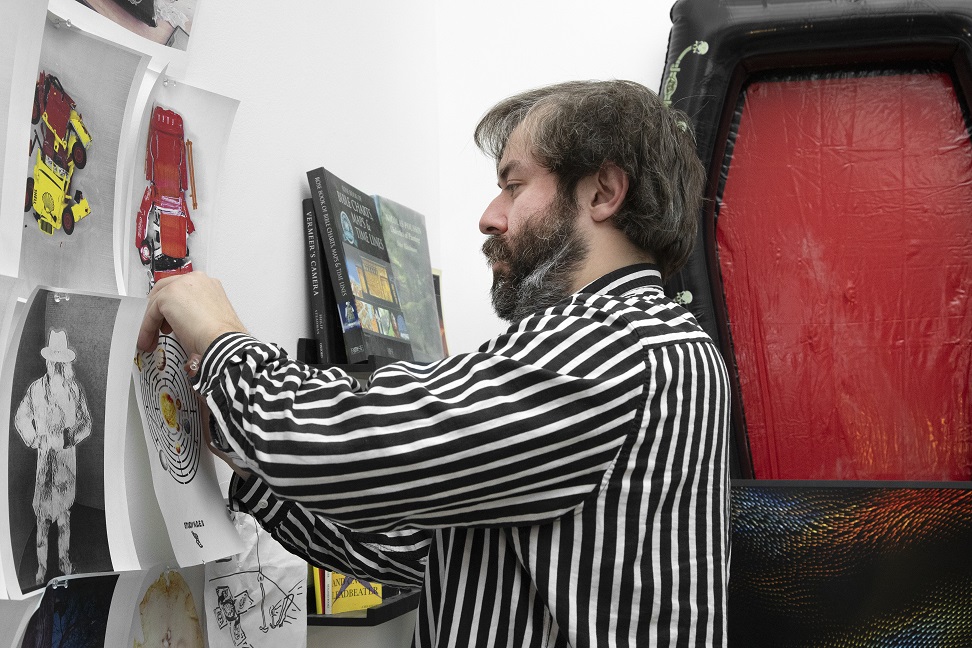 A person in a striped shirt is arranging papers with photos, illustrations and designs on a wall. Books are displayed on a narrow shelf in the corner behind the person. A tall red found object sculpture is seen in the background.