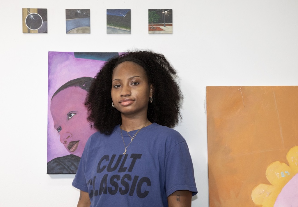An individual with curly hair stands in front of a wall displaying several artworks, including a painting with a person's face and an abstract piece. They are wearing a blue shirt with "Cult Classic" written on it in bold font.
