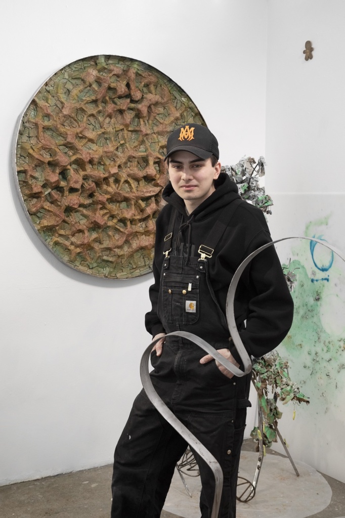 An individual stands in an art studio, wearing a black cap, overalls, and hoodie, in front of a textured circular artwork on the wall. There are sculptural pieces and a partially visible abstract artwork against the white walls.