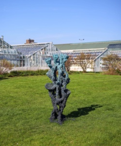 A large coral-like sculpture made from recycled plastic, dark gray at its base and transitioning to teal at the top, stands on a grass lawn in front of the glass greenhouse structures of Garfield Park Conservatory, Chicago. Photo: NaBeela Washington for 15 West.