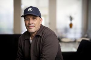 A person wearing a brown knit jacket and a dark colored baseball cap with a black and white "CA" logo on the front sits, slightly leaning forward, looking to the camera with a slight smile.